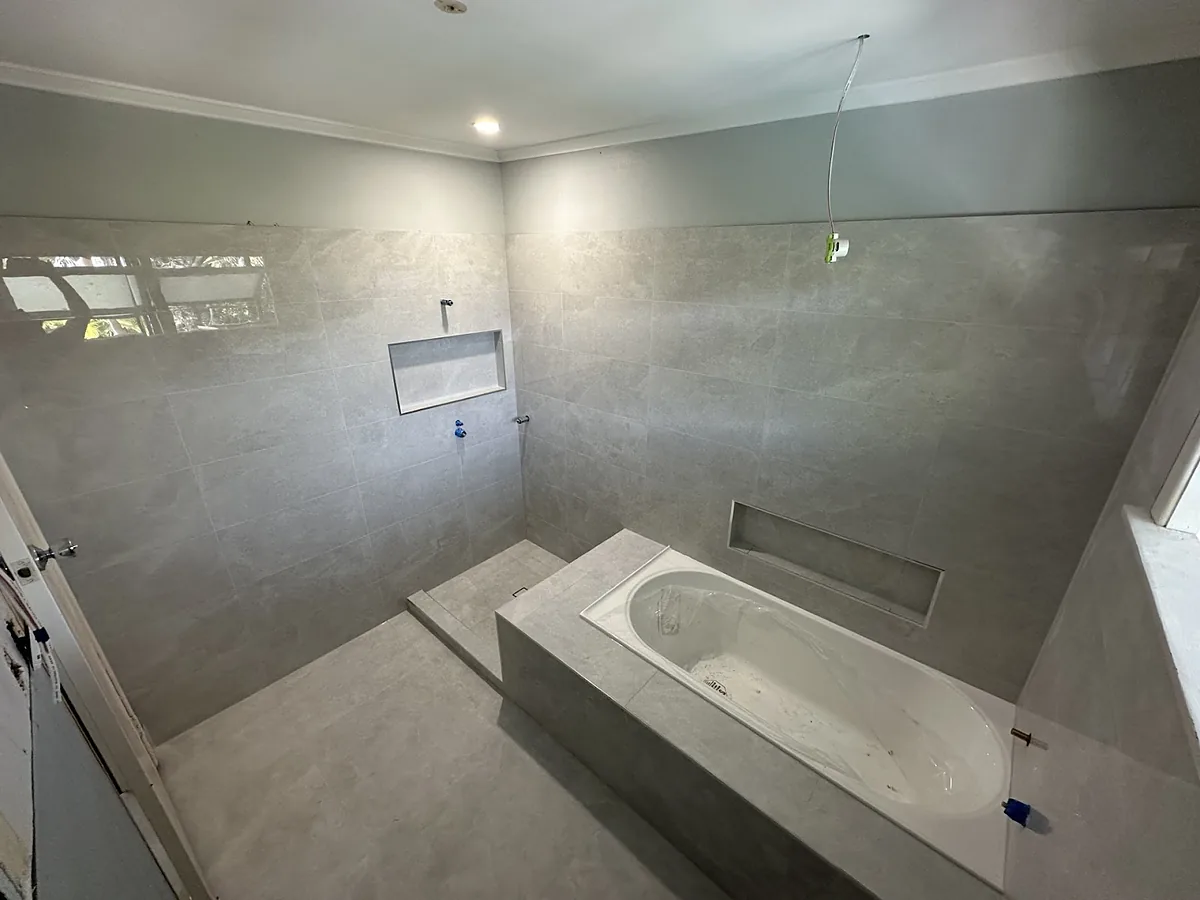 A renovated bathroom tiled floor to ceiling in pale grey, with a tiled bath hob, two niches and a step into the shower