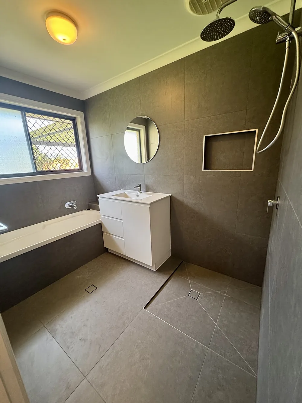 A dark grey bathroom with large format tiles, a round mirror, a framed niche and an envelope-cut shower floor