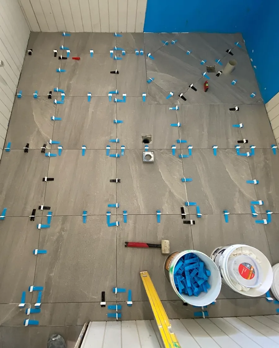 Large format grey floor tiles being laid with blue levelling clips and wedges, a rubber mallet and a spirit level