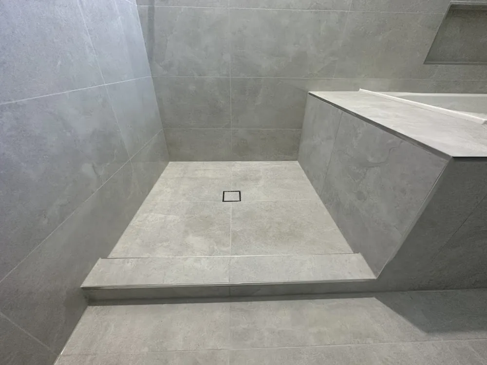 A finished walk-in shower in large grey tiles with a square drain, a tiled step and a tiled bench with metal trim
